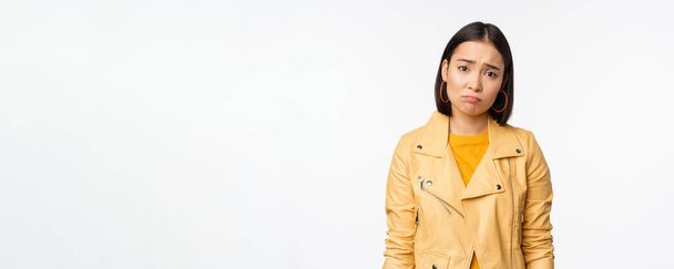 Портрет грустной корейской женщины, дующейся, хмурящейся и выглядящей расстроенной, расстроенное выражение лица, стоящей мрачно на белом фоне - Фото, изображение