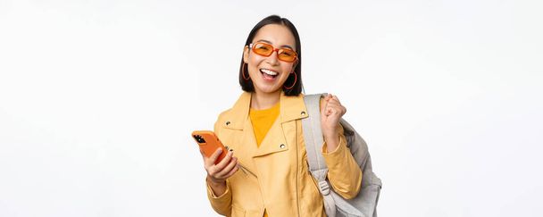 Joven mujer asiática viajero, chica turista en gafas de sol, la celebración de la mochila y el teléfono móvil, el uso de la aplicación en el teléfono inteligente, de pie sobre fondo blanco - Foto, imagen