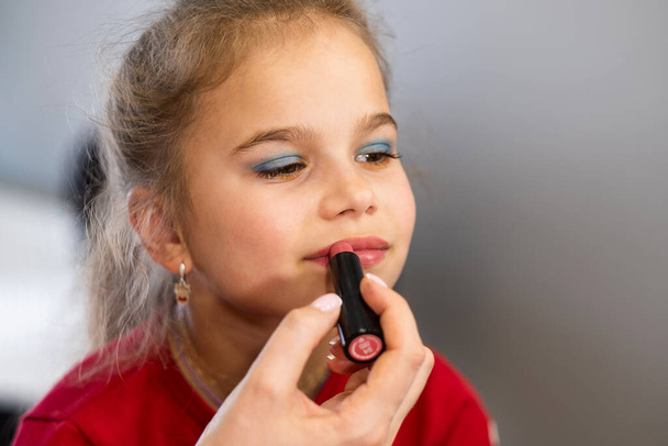 De make-up kunstenaar schildert de lippen van een klein meisje in de salon. Portret van een schattig meisje. - Foto, afbeelding