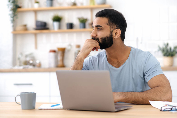 Thoughtful handsome indian or arabian man in casual t-shirt, freelancer, designer, working from home, sitting in the kitchen with a laptop, looking away, thinking about a new project, strategy - Фото, изображение