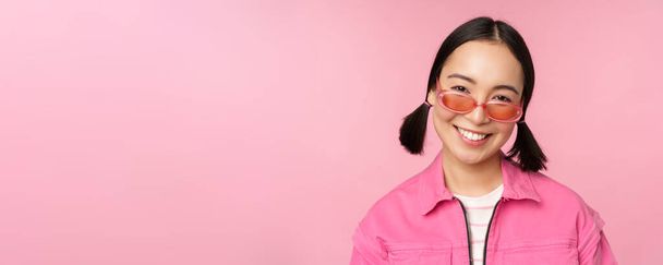 Close up of stylish korean girl in sunglasses, smiling happy, posing against pink background. People face concept - Foto, Imagen