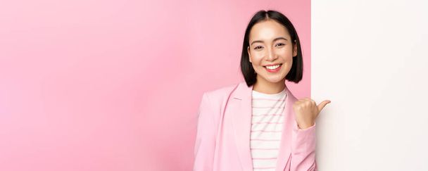 Portré mosolygó ázsiai üzletasszony öltönyben, vállalati hölgy ujjal mutogató fehér üres fal, fórumon info vagy reklám, álló felett rózsaszín háttér - Fotó, kép