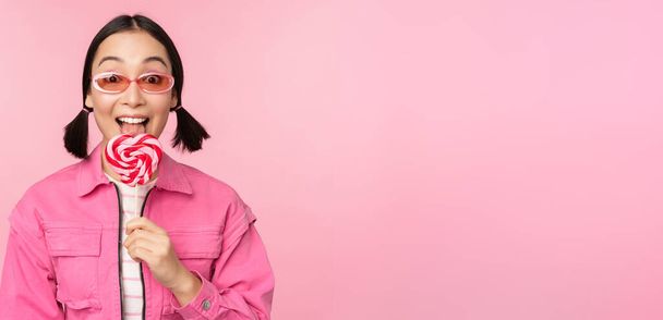 Élégant coréen fille lécher lolipop, manger des bonbons et sourire, debout dans des lunettes de soleil sur fond rose - Photo, image
