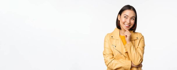 Portrait of stylish korean woman in yellow jacket, smiling thoughtful, thinking and looking up at logo or advertisement, standing over white background - Foto, afbeelding
