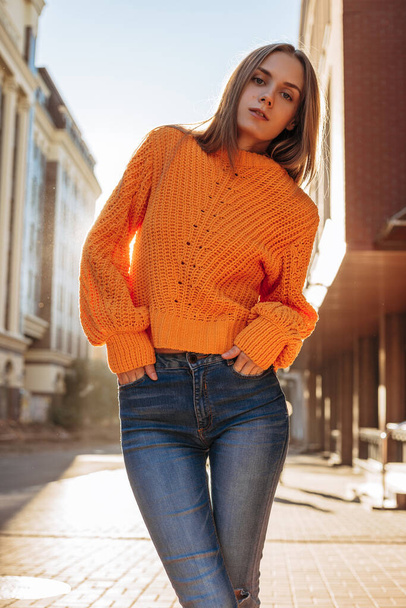 Medium full shot portrait of the young blonde woman on the city street background in the sunset. The girl wearing red knitted sweater posing at the camera - Foto, imagen