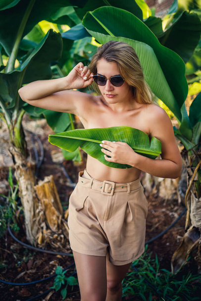 A beautiful young woman stands in a beautiful pose near a banana tree, her little girl covers her breasts with a leaf on a banana plantation. - Foto, Imagem