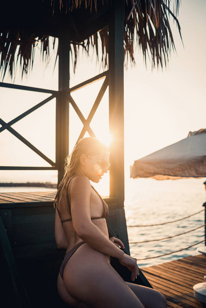 Attractive young woman in a beige bikini swimsuit at sunset sits in a wooden house by the sea - 写真・画像