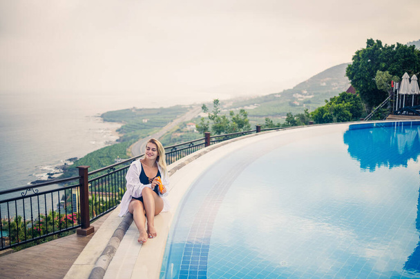 Mooie aantrekkelijke jonge vrouw zit in de buurt van een groot zwembad met uitzicht op de zee. Vakantie concept. Meisje in een zwart badpak smeert haar huid met zonnebrandcrème - Foto, afbeelding