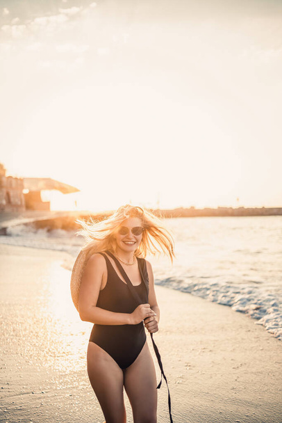 Young beautiful woman in a black swimsuit and hat with glasses walks along the beach in Turkey at sunset. The concept of sea recreation. Selective focus - Photo, Image