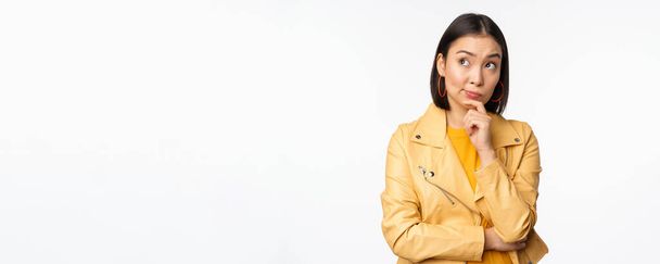 Portret van stijlvolle Koreaanse vrouw in geel jasje, lachend bedachtzaam, denkend en kijkend naar logo of advertentie, staand over witte achtergrond - Foto, afbeelding