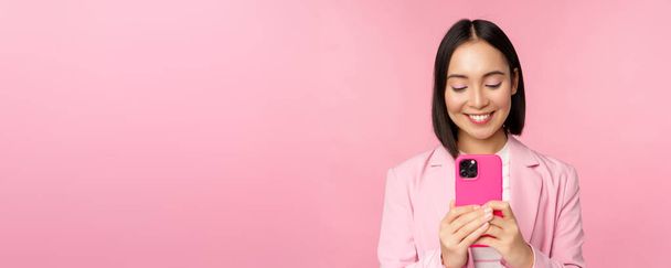 Портрет усміхненої ділової жінки, азіатської корпоративної людини, що використовує смартфон, мобільний додаток, стоїть на рожевому фоні
 - Фото, зображення