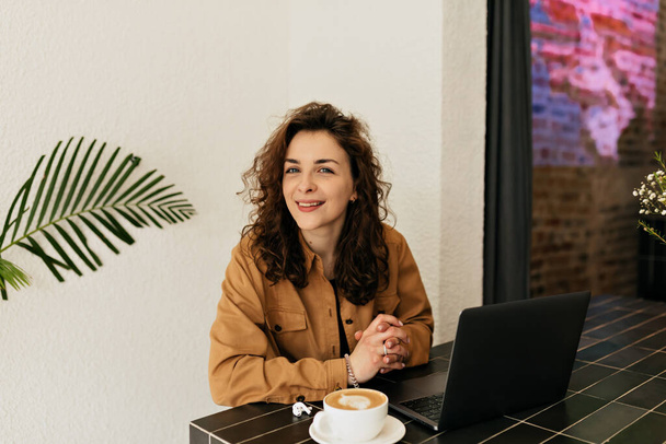 Charmantes Mädchen mit dunklem lockigem Haar mit Lächeln blickt in die Kamera, posiert am Tisch im Café mit Cappuccino und arbeitet am Laptop. - Foto, Bild