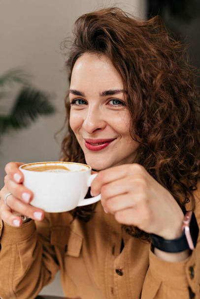 Detailní fotografie z interiéru okouzlující usměvavá dáma drží šálek cappuccino a usmívá se. Rozkošná hezká žena s kudrlinkami a krásný úsměv těší ranní kávu - Fotografie, Obrázek