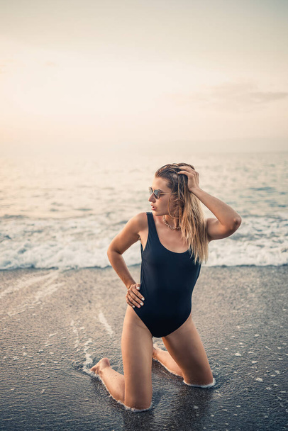 Attractive young woman in sunglasses kneels in the sea at sunset light in a black swimsuit. Selective focus - Zdjęcie, obraz
