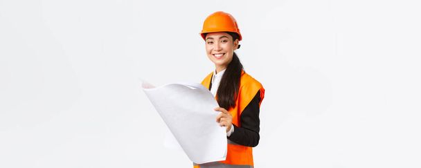 Pleased smiling female asian construction manager following blueprints during building house, looking satisfied at camera, studying architect plan, standing white background in safety helmet - Photo, Image