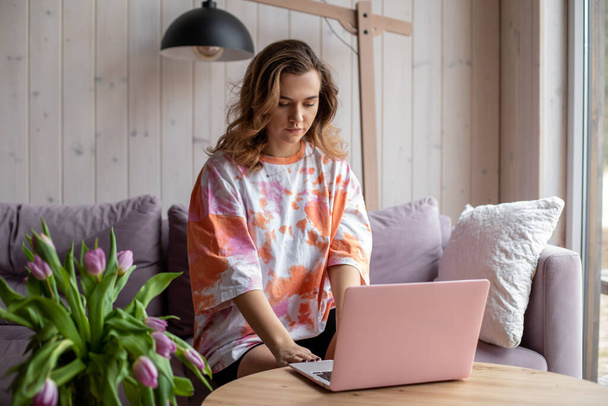 Portrait of woman typing on modern pink laptop and sitting on soft sofa in casual clothes near table with vase of fading tulips. Living room in elegant design with aesthetic decor, free copy space - Fotografie, Obrázek