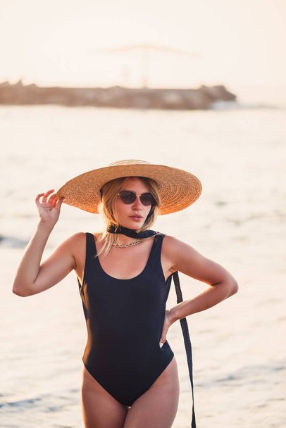 Beautiful girl in a black swimsuit and hat on a sandy beach at the sea in the sunset sunlight - Photo, Image