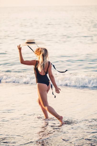Όμορφο κορίτσι με μαύρο μαγιό και καπέλο σε μια αμμώδη παραλία στη θάλασσα στο ηλιοβασίλεμα. Επιλεκτική εστίαση - Φωτογραφία, εικόνα