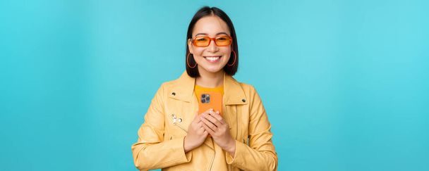 Obraz stylowej, pięknej Chinki w okularach przeciwsłonecznych, trzymającej smartfona, robiącej zdjęcie na telefonie komórkowym i uśmiechniętej, nagrywającej smth, stojącej nad niebieskim tłem - Zdjęcie, obraz