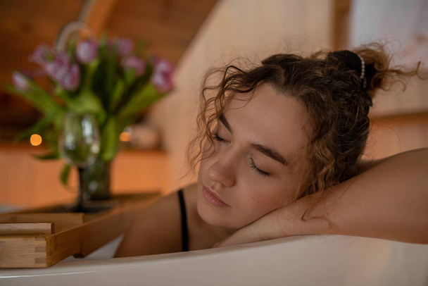 Portrait closeup of naked woman with closed eyes relaxing and spending weekend at home, blurred background. Taking bath with glass of wine on food tray and bouquet of tulips in cozy wooden bathroom - Photo, Image