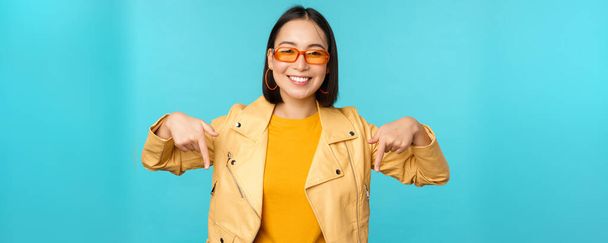 スタイリッシュなアジアの女の子の肖像画、サングラスをかけ、笑顔とポイントの指を下に、広告を示し、青い背景の上に立つ - 写真・画像