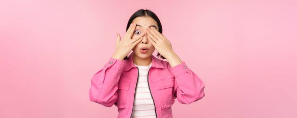 Porträt eines asiatischen Mädchens guckt aufgeregt durch Finger, bedeckt Augen, sieht Überraschung, steht über rosa Hintergrund - Foto, Bild
