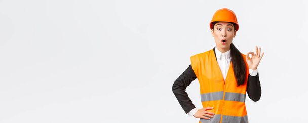 Vestido y sorprendido asiático gerente de construcción femenina satisfecho con la calidad increíble, de pie en el casco de seguridad y chaqueta reflectante, mostrando gesto bien y decir wow, fondo blanco
 - Foto, imagen