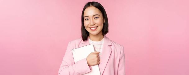 Imagen de una joven mujer corporativa coreana, directora general sosteniendo una tableta digital, sonriendo y luciendo profesional, usando traje, de pie sobre fondo rosa - Foto, imagen