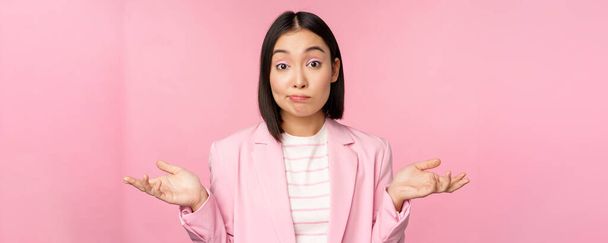 Portrait of confused asian businesswoman shrugging shoulders, looking clueless and puzzled, dont know, cant say, standing over pink background in office suit - Photo, Image