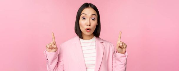 Portré ázsiai üzletasszony, vállalati dolgozó öltöny, úgy néz ki, meglepte a reklám, mutat ujj fel, mutatja banner vagy logó a tetején, áll át rózsaszín háttér - Fotó, kép