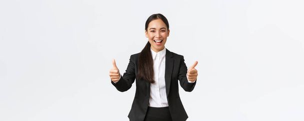 Successful upbeat asian businesswoman showing thumbs-up and smiling happy. Cheerful lady manager in business suit rejoicing ofer good news, say nice work, well done or congrats gesture - Photo, image