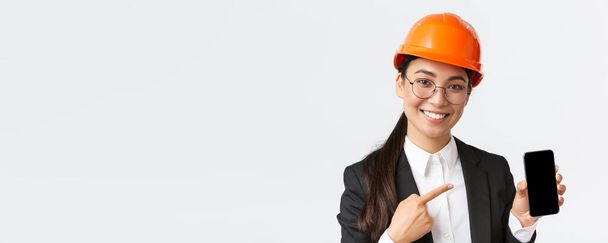 Detailní záběr usměvavé profesionální ženské asijské stavební inženýrky, architektky v obleku a bezpečnostní masce představit aplikaci, ukazující prst na displej smartphonu, bílé pozadí - Fotografie, Obrázek