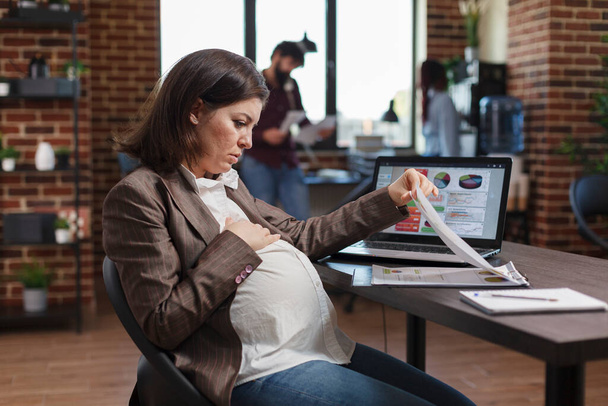 Zwangere vrouw ondervraagt data grafieken en business analytics resultaten tijdens het zitten aan het bureau. - Foto, afbeelding