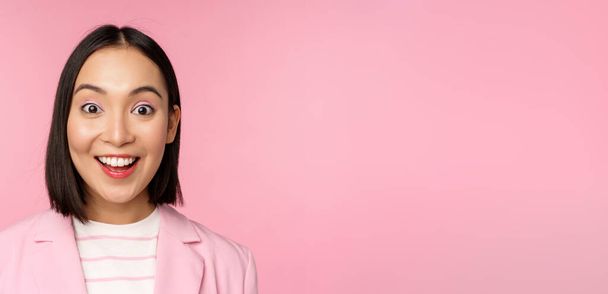 Close up portrait of asian corporate woman, business lady looking surprised and amazed at camera, standing in suit against pink background - Photo, Image
