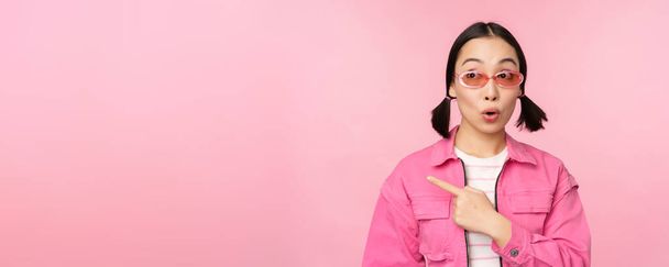 Ritratto di ragazza asiatica sorridente in abito elegante, occhiali da sole, indicando il dito sinistro, mostrando pubblicità, banner, in piedi su sfondo rosa - Foto, immagini