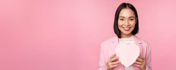 Це для тебе. Романтична мила азіатська корпоративна жінка, дівчина в костюмі, показує коробку у формі серця з подарунком, що стоїть на рожевому фоні
 - Фото, зображення