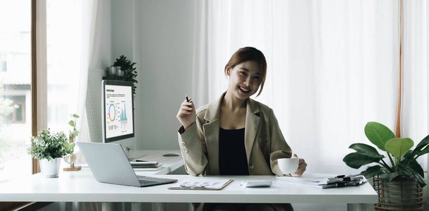 Посміхнена азіатська бізнесменка, що тримає чашку кави і лептоп в офісі. Дивлячись на камеру - Фото, зображення