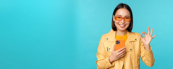 glimlachend aziatisch meisje met smartphone, shwoing oke, ok teken in goedkeuring, staande over blauwe achtergrond - Foto, afbeelding