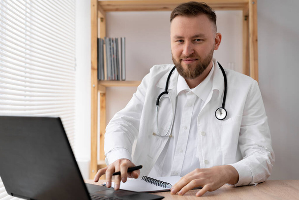 Porträt eines jungen Arztes im weißen Kittel mit Stethoskop, der in einer Arztpraxis sitzt und am Laptop arbeitet. - Foto, Bild