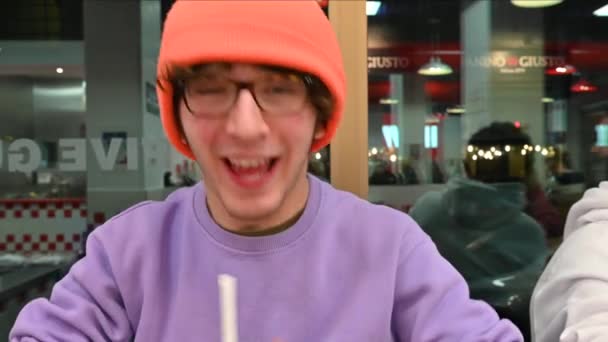 Portret van een schattige blanke jongeman met een bril en een oranje pet: hij praat en grapt voor de camera terwijl hij wat drinkt in een club. - Video