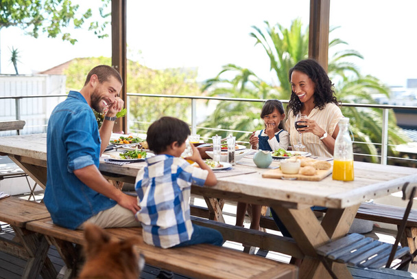 Meals always taste better when enjoyed with family. Shot of a beautiful young family enjoying themselves while having a meal together around a table outdoors. - Foto, Imagen