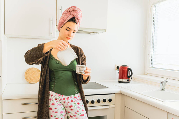 teenage girl with bath towel on wet hair in pajamas stands in kitchen, pours milk from milk jug into bowl with fragrant morning coffee. Sunday is day off - 写真・画像