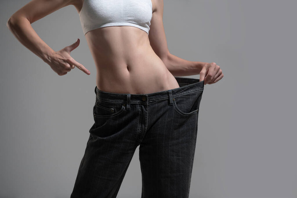 Dieting concept with oversized jeans. Skinny woman in too large jeans. Concept of successful weight loss. Thin girl in big trousers on a gray background. - Photo, Image