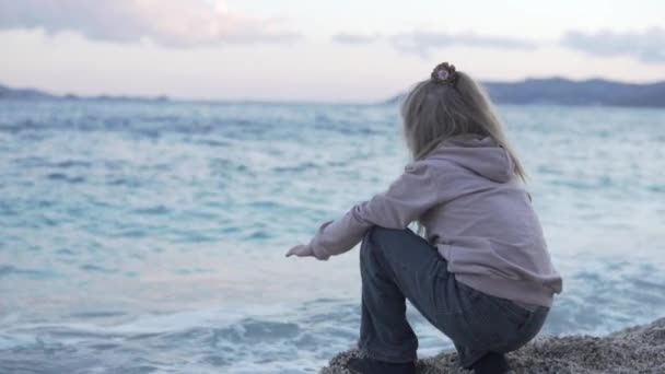 Un bambino si siede su una spiaggia di ciottoli vicino al mare - Filmati, video