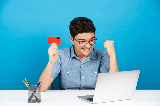 Feliz tipo hispano celebrando la compra en línea. Adolescente celebración de la tarjeta de crédito en frente de la computadora portátil aislado sobre fondo azul. - Foto, imagen