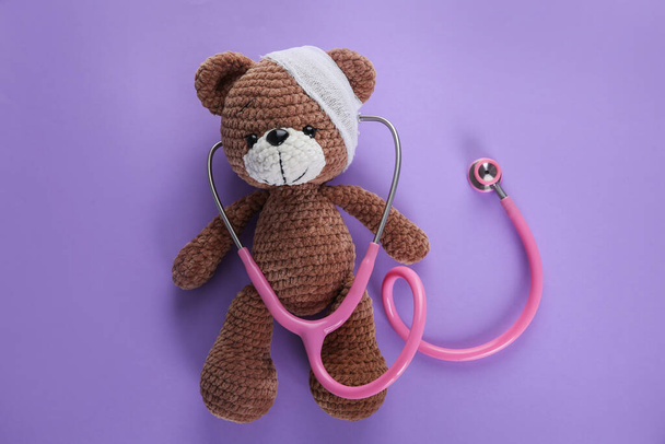 Cute toy bear with bandage and stethoscope on violet background, top view. Children's hospital - Φωτογραφία, εικόνα