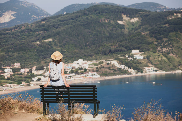 κορίτσι τουρίστρια κάθεται σε ένα παγκάκι στο κατάστρωμα παρατήρησης και κοιτάζει τη θάλασσα και ένα όμορφο τοπίο, Κέρκυρα στην Ελλάδαη θάλασσα - Φωτογραφία, εικόνα