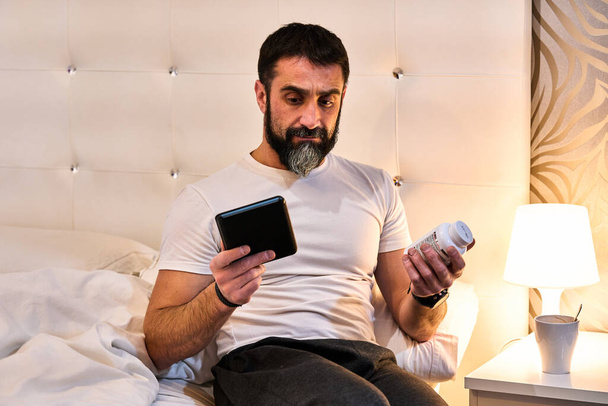 άνθρωπος που δεν μπορεί να κοιμηθεί με αμφιβολίες μεταξύ της ανάγνωσης ενός ebook ή τη λήψη του φαρμάκου που έχει σε ένα μπουκάλι χάπια - Φωτογραφία, εικόνα