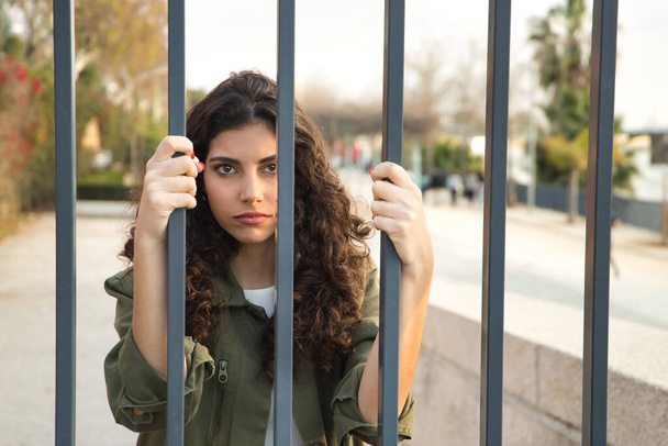 Portrait of a young and beautiful woman, brunette, with curly hair, with a green jacket and white shirt, clinging to bars with a serious look. Concept beauty, fashion, trend, model, seriousness. - Photo, Image