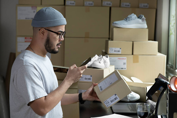 Asian male small business owner using mobile app on smartphone checking parcel box. Warehouse worker, seller holding phone scanning retail drop shipping package postal parcel bar code. - Foto, Imagen